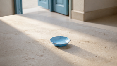 A broad shaft of window light falling across honed pale limestone, with one empty pale blue glazed ceramic dish standing in the light and casting a long soft shadow.