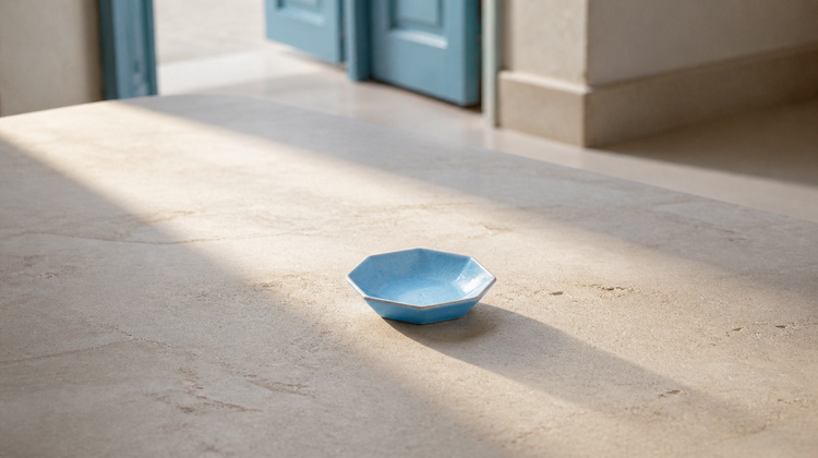 A broad shaft of window light falling across honed pale limestone, with one empty pale blue glazed ceramic dish standing in the light and casting a long soft shadow.