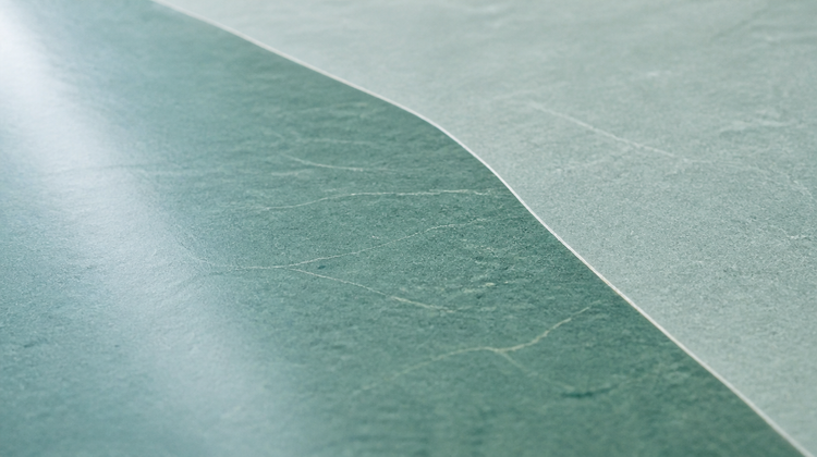 A honed sea-green stone slab half covered by a thin, perfectly clear film of liquid, its glossy wet sheen meeting the matt dry stone along one clean curving edge