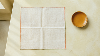 An overhead flat-lay of a square of ivory cotton muslin with a terracotta stitched border on pale lime plaster, beside an empty amber-glazed ceramic dish - a calm image for an article on breastfeeding-safe skincare.