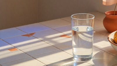 A clear glass of plain drinking water on a sunlit tiled kitchen counter beside a terracotta pot and fresh fruit