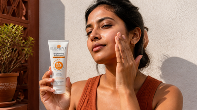 A young Indian woman applies The Element Brightening Sunscreen SPF 50+ to her cheek in strong daylight on a sunlit terrace.