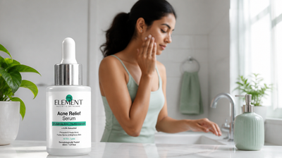 An Indian woman in a bright bathroom beside The Element Acne Relief Serum with 2% salicylic acid and 5% niacinamide.
