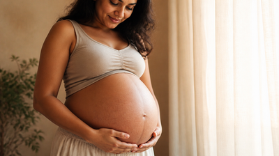 A pregnant Indian woman cradles her bump in soft window light, natural skin texture visible.