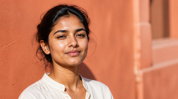 Indian woman outdoors in strong sunlight against a terracotta wall, skin carrying the fresh sheen of just-applied sunscreen
