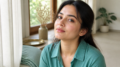 Indian woman with clear, even skin sitting in daylight at a home window, wearing a teal-green shirt