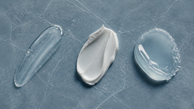 Three skincare textures compared on blue-grey slate: a clear humectant gel, a matte emollient cream and a glossy occlusive balm