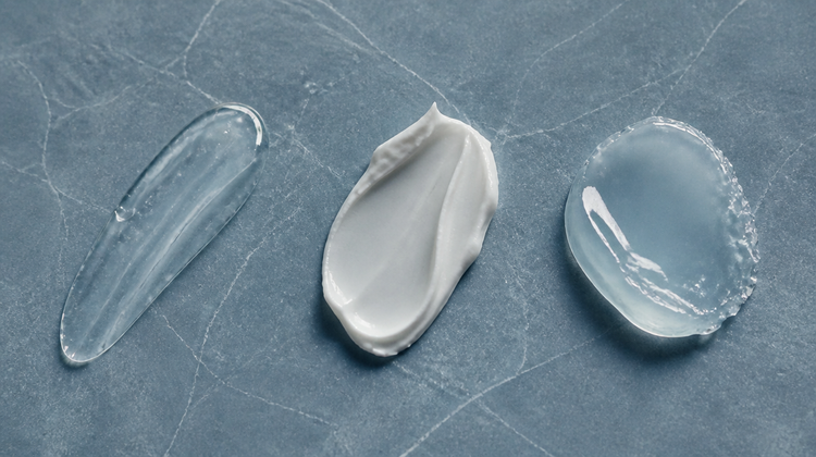 Three skincare textures compared on blue-grey slate: a clear humectant gel, a matte emollient cream and a glossy occlusive balm