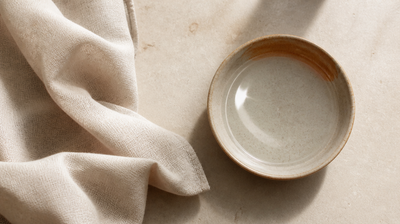 Overhead still life of folded raw linen beside a handmade ceramic dish of clear water on honed stone