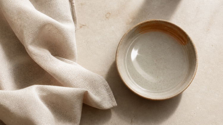 Overhead still life of folded raw linen beside a handmade ceramic dish of clear water on honed stone