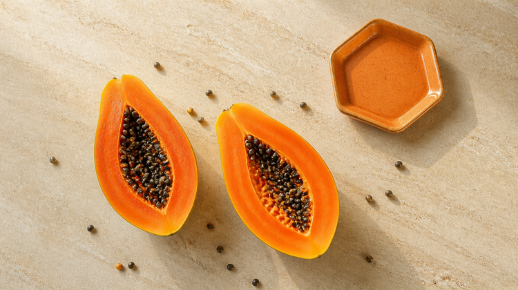 A ripe papaya halved lengthways on pale cream stone with glossy dark seeds, shot from directly overhead beside a terracotta dish