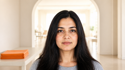 A woman with soft diffuse uneven pigmentation on her upper cheeks standing in a bright daylit room, an archway opening behind her, with folded amber-orange linen on a low table beside her