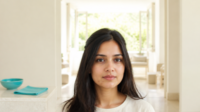 A woman standing calmly away from the mirror in a bright daylit room, hands out of frame, with a turquoise glazed bowl and folded turquoise linen on a stone plinth beside her