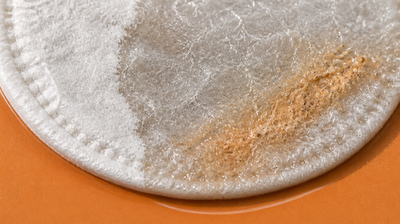 Macro view of a round cotton pad on a warm orange tile, dry and fluffy on one side, soaked and matted on the other, with a beige smudge of lifted grime sitting on the wet fibres.