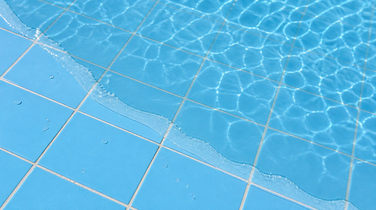 Close view of the edge of clear water on blue pool tiling, sunlight caustics rippling across the submerged tiles and dry matt tile beyond the receding waterline.