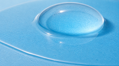Macro of two clear liquid layers on a sky-blue tile: a thin continuous film across the surface with a domed lens of gel resting on top of it, the two merging seamlessly.