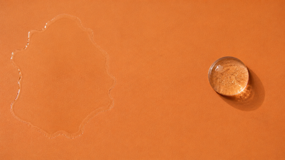 Two pools of clear liquid on a warm orange tile: a thin watery one spread wide and drying at its edges, beside a single thick domed bead of gel holding its shape.