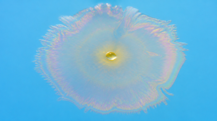 A single drop of aromatic oil spreading across water into one thin iridescent film, its edge feathering into pale gold, rose and green interference bands on a sky-blue ground.
