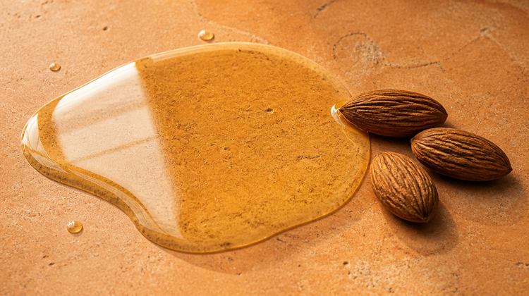 A clear golden pool of sweet almond oil spreading across honed terracotta stone beside three whole almonds