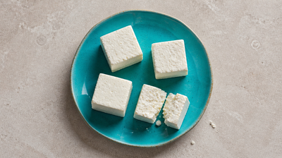 Knife-cut cubes of fresh Indian paneer on a teal ceramic plate, one broken open to show its crumbly white interior