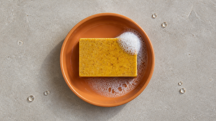 A speckled turmeric soap bar with white lather in a terracotta dish, beside water droplets on stone