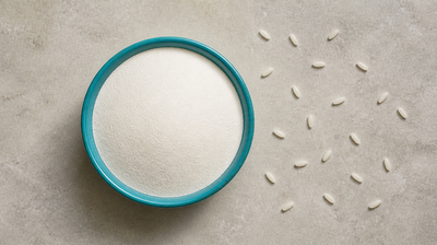 A bowl of fine white refined wheat flour beside scattered raw white rice grains on pale stone