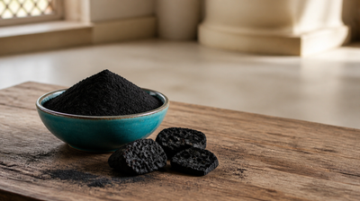 Matte black activated charcoal powder heaped in a teal ceramic bowl beside porous charcoal chunks on aged teak