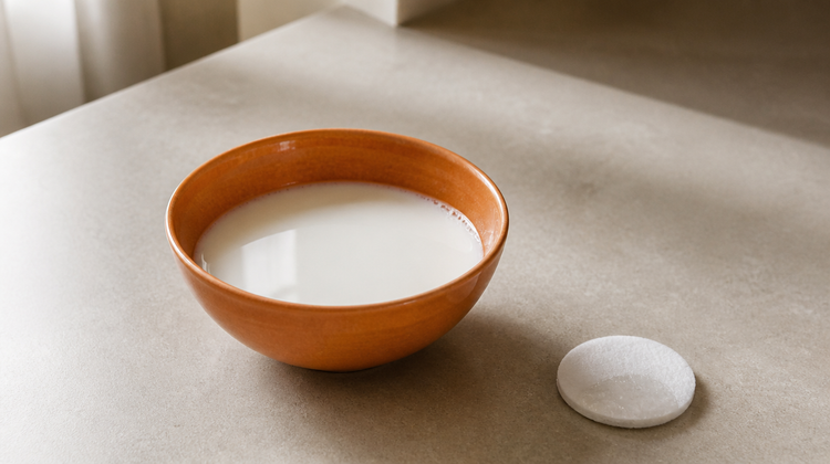 Fresh raw milk in an orange-glazed ceramic bowl on pale stone, with a cotton pad beside it
