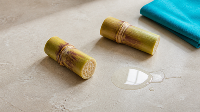 Two cut lengths of sugarcane, the source of glycolic acid, beside a shallow pool of clear viscous liquid on pale grey honed stone with a folded teal cloth.