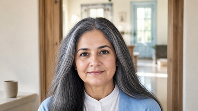 An Indian woman in her early sixties with silver-threaded hair, standing in a bright home doorway in a pale blue cardigan and white collared shirt.