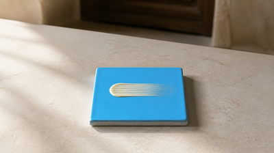 A short matt swipe of pale yellow cream dragged across a small sky-blue glazed ceramic tile on pale grey stone, thinning to a sheer veil.