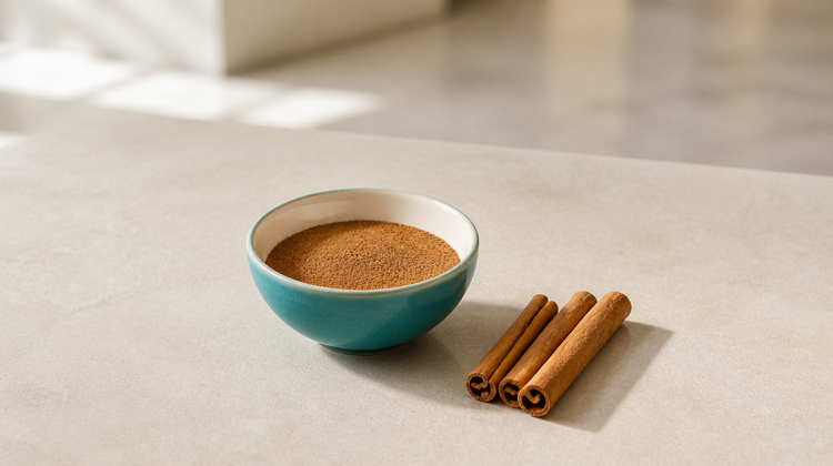 Teal ceramic bowl of ground cinnamon powder with three cinnamon sticks on pale stone