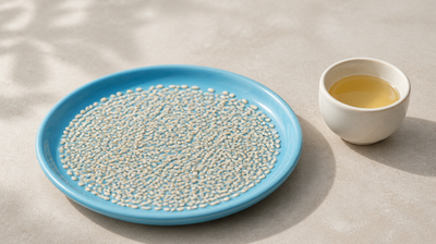 Sky-blue ceramic saucer scattered with white sesame seeds beside a small cup of pale golden sesame oil