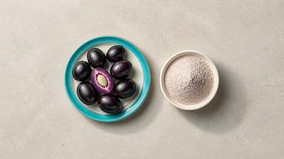 Ripe jamun fruits and a halved jamun showing its seed on a teal plate, beside a bowl of jamun seed powder