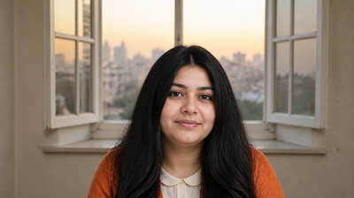 A young Indian woman in a burnt-orange cardigan at home at dawn after a night shift, city window behind her