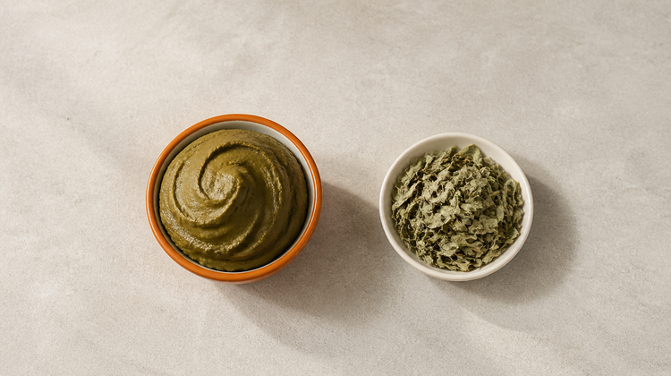 A burnt-orange bowl of freshly mixed natural green henna paste beside a dish of dried henna leaves, seen from above