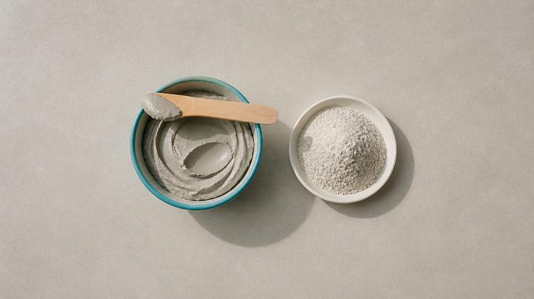 Overhead view of a teal bowl of grey bentonite clay mask paste with a wooden spatula and a dish of dry clay powder