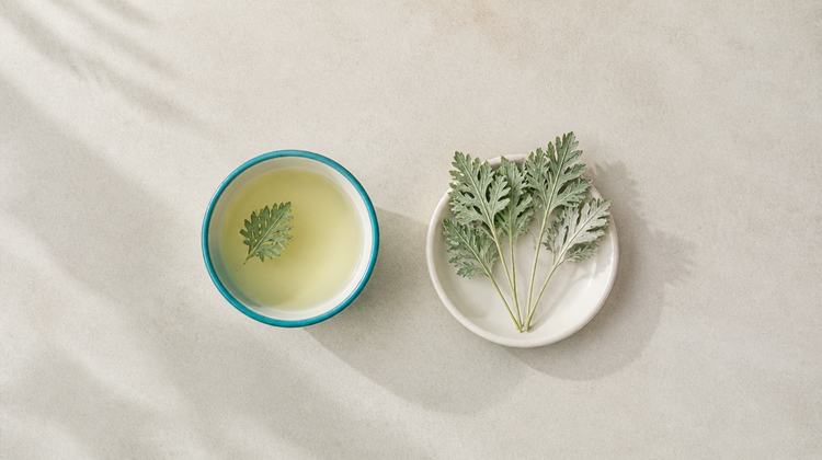 Pale mugwort leaf water in a teal bowl beside fresh sage-green mugwort sprigs, seen from above