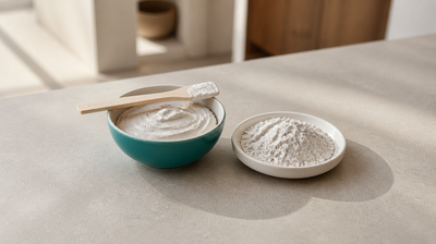 Teal bowl of matt white kaolin clay mask paste with a wooden spatula beside a dish of white kaolin clay powder