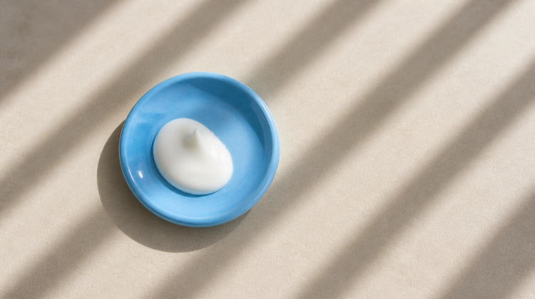A dollop of white sunscreen lotion in a sky-blue ceramic saucer under bright window sunlight, for a guide to Tinosorb S and M UV filters