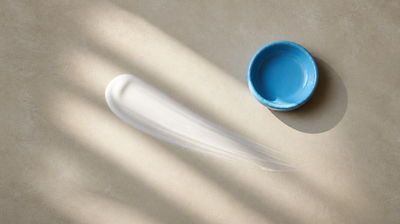 A swept stroke of white sunscreen cream beside a sky-blue dish in bands of sunlight, illustrating the UVA filter Uvinul A Plus