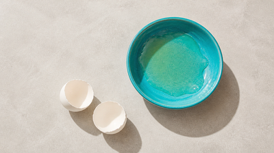 Raw separated egg white in a teal ceramic bowl beside two empty eggshell halves on pale stone