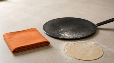 Black iron tawa, rolled chapati and flour beside a folded orange kitchen cloth on a stone counter