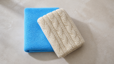 A folded sky-blue cotton towel and a cream cable-knit wool throw on pale limestone, for an article on dry legs in winter