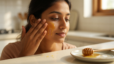 Indian woman applying a thin layer of raw honey to her cheek at a kitchen counter, with a wooden honey dipper in a ceramic dish