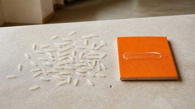 Raw long-grain rice scattered on pale stone beside a clear serum swipe on an orange tile, comparing kojic acid and niacinamide