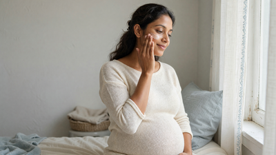 A pregnant Indian woman applies moisturiser to her cheek in soft daylight at home.