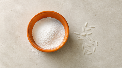 An orange ceramic bowl of fine white rice flour beside a scatter of long-grain basmati rice on pale limestone