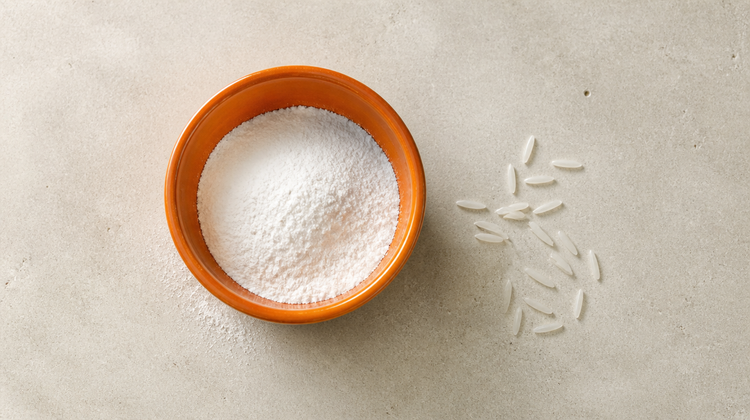 An orange ceramic bowl of fine white rice flour beside a scatter of long-grain basmati rice on pale limestone