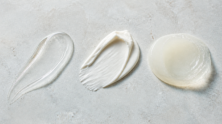 Three skincare textures on warm pale stone: a clear serum, a whipped cream and a translucent occlusive balm, the layers of a daily routine